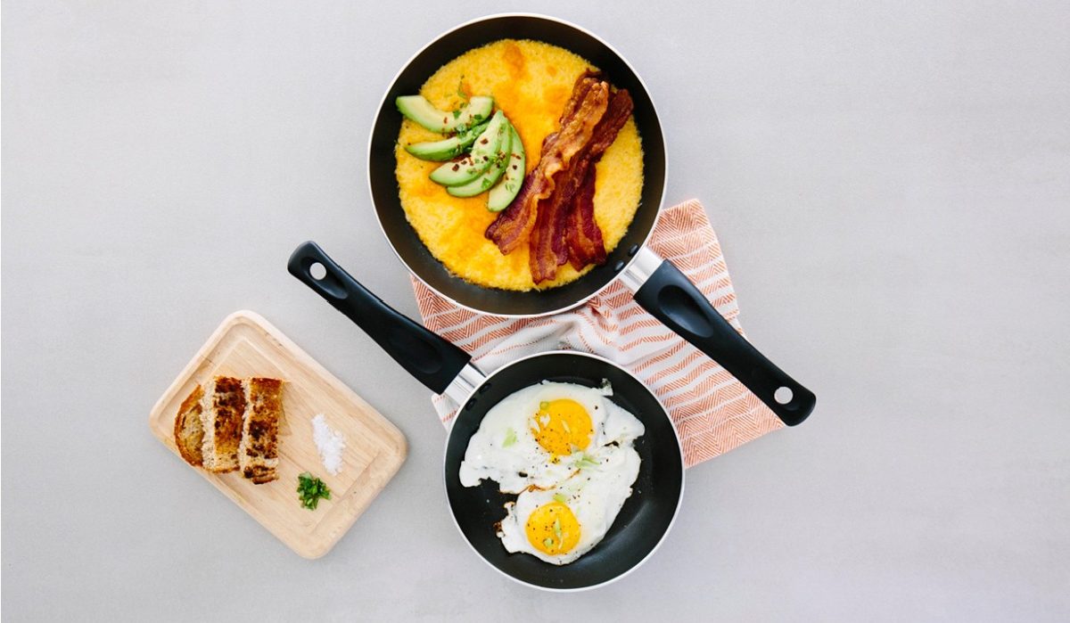two frying pans with eggs and bacon, on a towel, next to a cutting board with bread