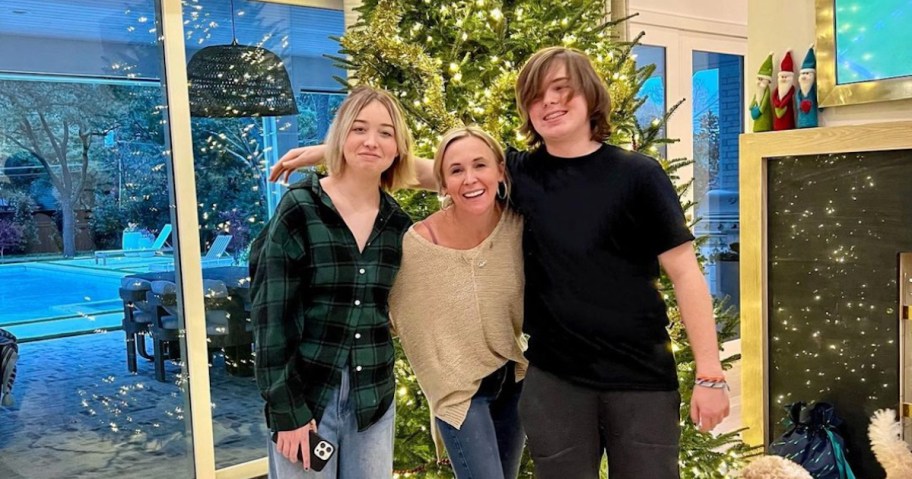 woman and two teens standing with arms around each other in front of lit christmas tree