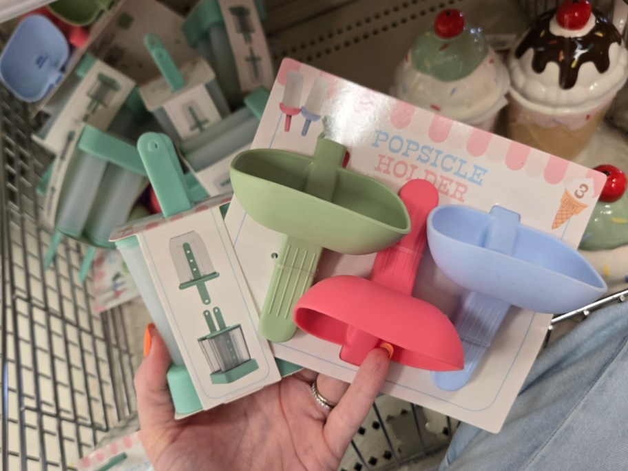 A hand holds a package of colorful popsicle holders in green, red, and blue, labeled "Popsicle Holder," amidst more holders on a store shelf.