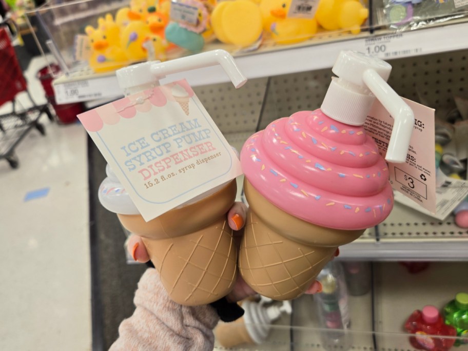 Two ice cream cone-shaped syrup pump dispensers with pink tops and sprinkles are held in a store. The tag reads "Ice Cream Syrup Pump Dispenser."