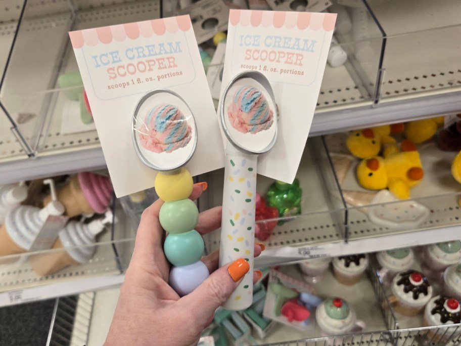 Two ice cream scoopers with pastel handles, one with round beads and the other with sprinkles. Background shows colorful novelty items on shelves.