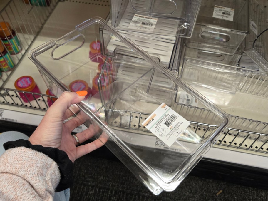 A hand holds a clear plastic storage container labeled with a price and barcode, in a store aisle with similar containers and colorful jars nearby.