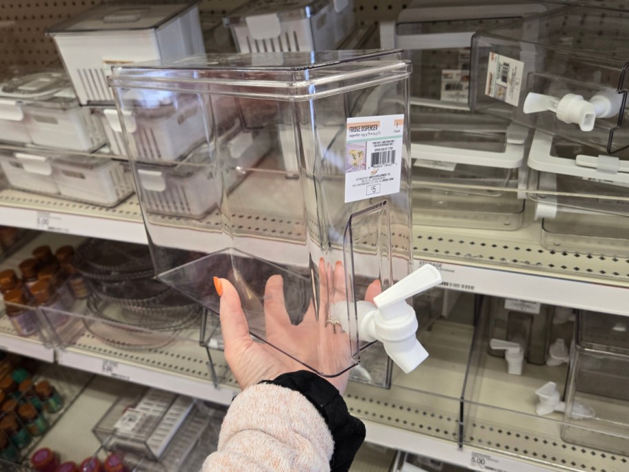 A hand holds a clear plastic beverage dispenser with a white spigot in a store aisle. Shelves display more dispensers and storage containers.