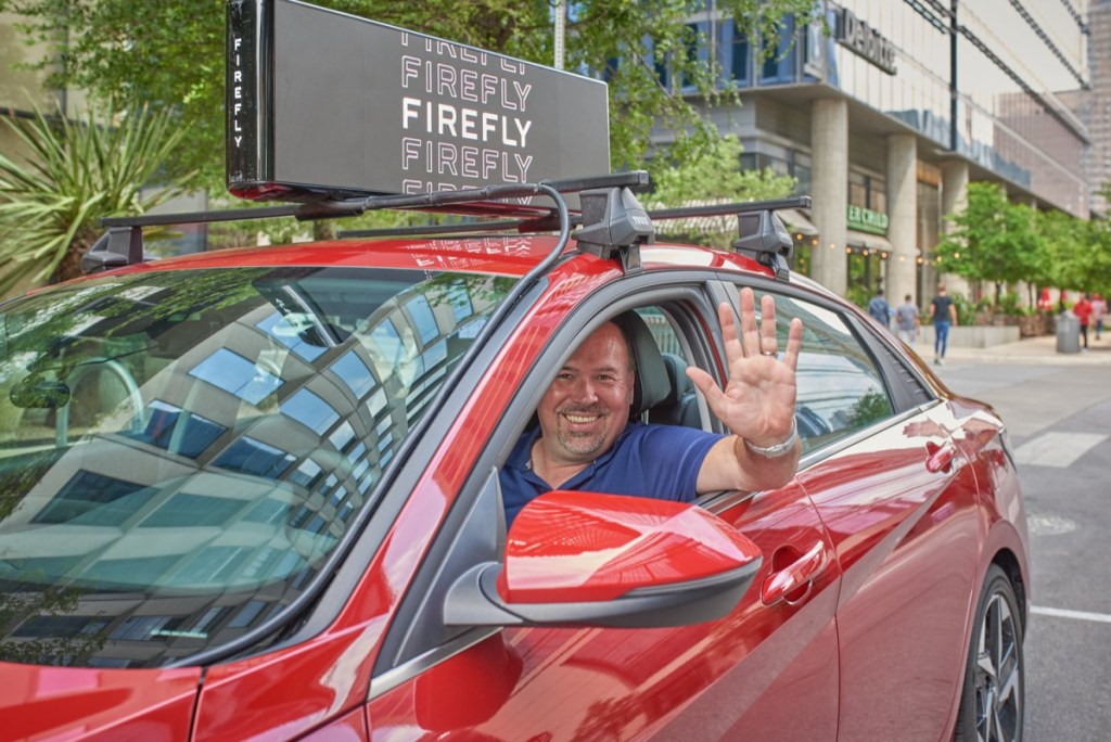 A man driving a car with a Firefly car advertisement on the roof