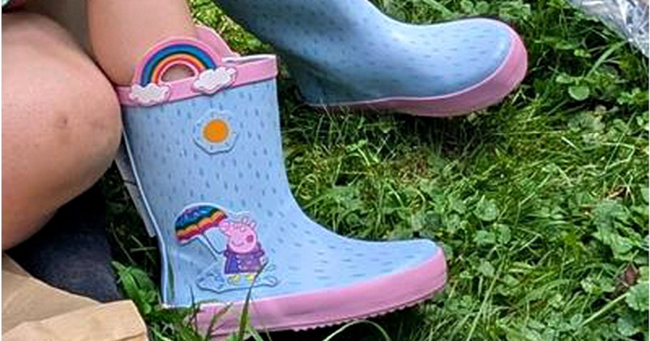 Children's blue rain boots with pink soles featuring colorful rainbows and a Peppa Pig holding an umbrella, set on vibrant green grass.