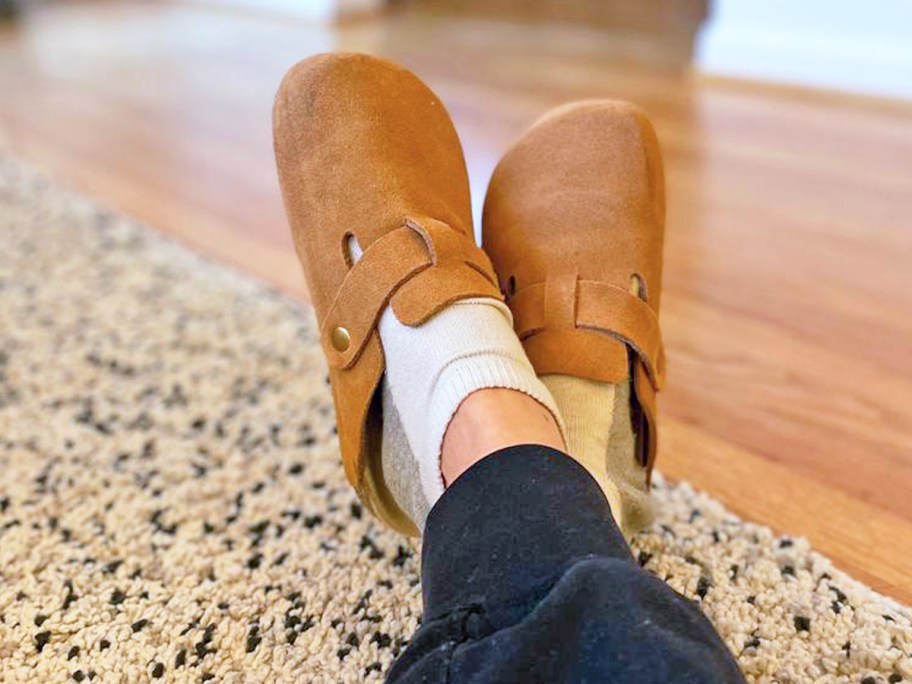 feet propped up and crossed wearing a brown color of suede clog mules with white socks