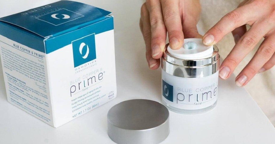 Hands press down on a jar of Blue Copper 5 Prime face cream beside its box.