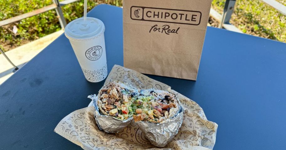 A Chipotle Burrito on a table next to a drink and a Chipotle bag