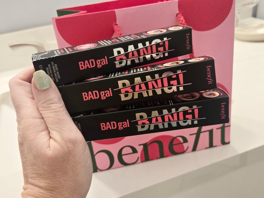 hand holding three benefit mascara boxes with gift bag behind it