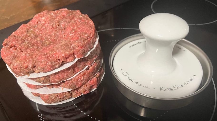 Stack of ground beef burgers next to burger patty press on stove top 
