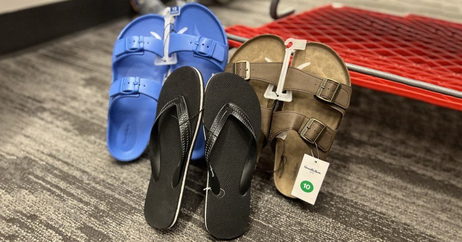 Three pairs of men&rsquo;s sandals on display at Target, including blue slide sandals with double buckles, classic black flip-flops, and tan Goodfellow & Co. sandals with dual buckle straps