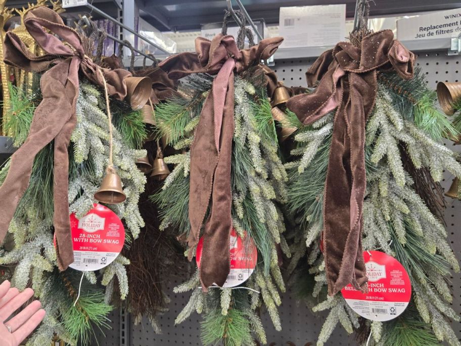 Holiday Time Bells, Bow, & Greenery Decorative Christmas Door Swags hanging in store