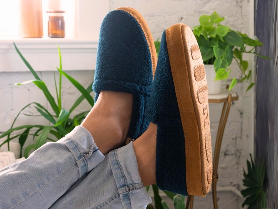 a mans' legs and feet propped up of the end of a sofa, he's wearing grey jogger pants and navy blue slippers with tan soles