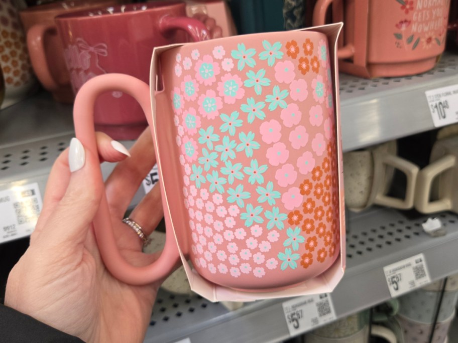 A hand holding a pink mug with a floral pattern of blue, pink, and orange flowers. Background shows more mugs on store shelves.