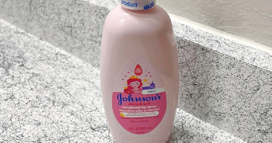 A pink bottle of Johnson's Kids Shiny & Soft Conditioning Spray on a marbled countertop. The label features a cartoon girl and a castle.