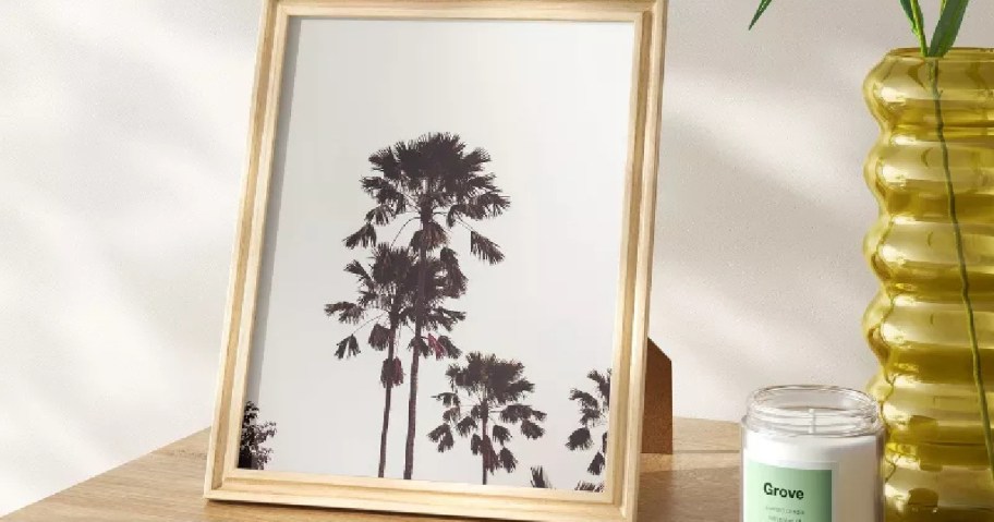 Framed photo of tall palm trees in silhouette with a light background, on a wooden table. Nearby, a yellow glass vase and a white candle labeled "Grove."