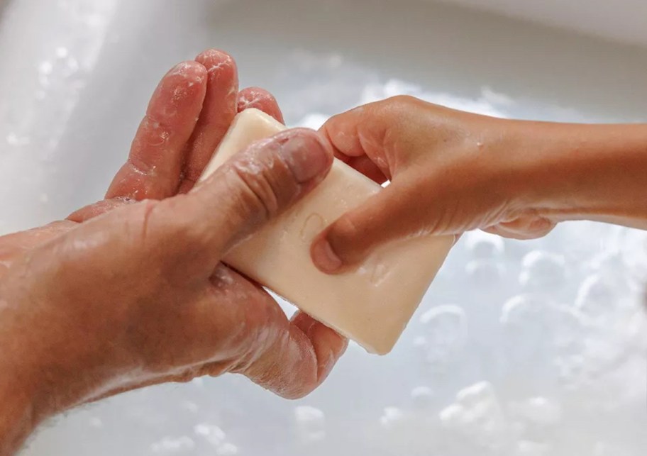 two hands of adult and child holding white bar of soap over water