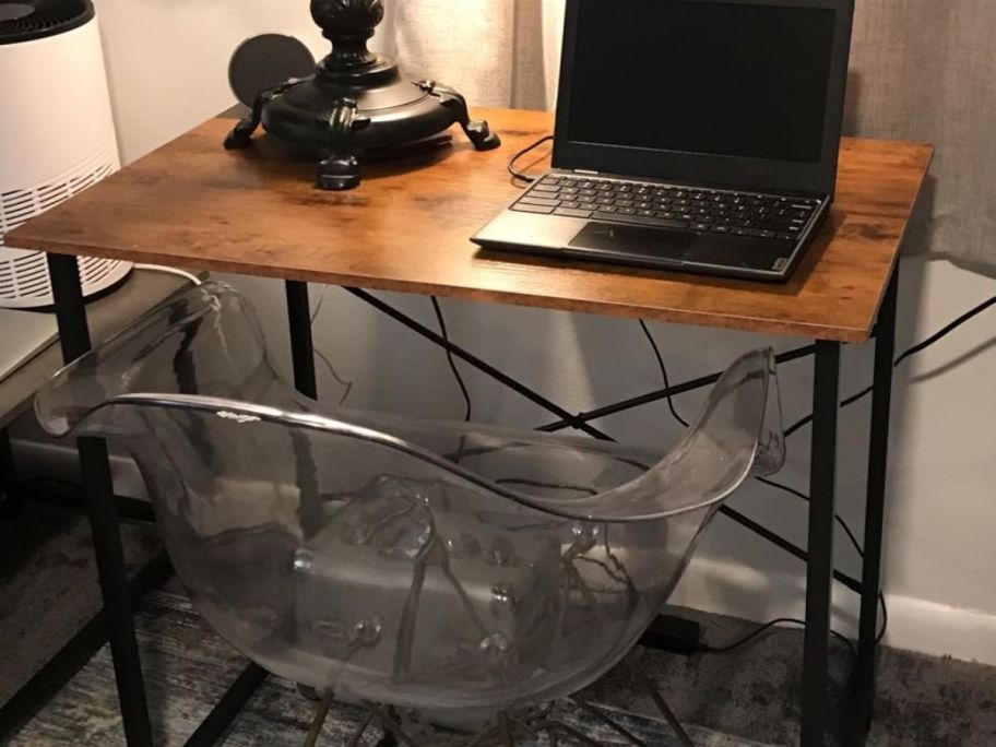A brown computer desk with black metal legs. It has a lamp and a laptop on it and a clear chair tucked under it. 