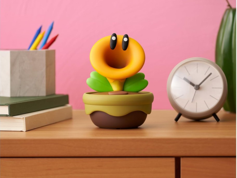 A whimsical toy plant with big eyes and a yellow trumpet-shaped top sits on a wooden desk between a stack of books and a clock, against a pink wall.