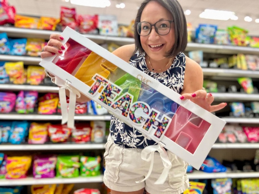 smiling woman standing in a store aisle holding a TEACH snackle box