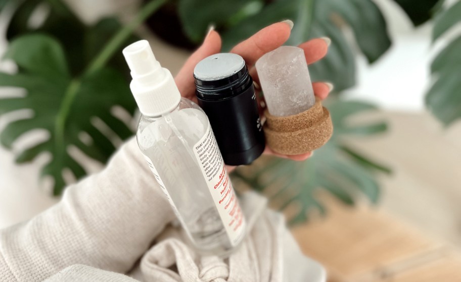 hand holding spray stick and crystal deodorants in front of monstera