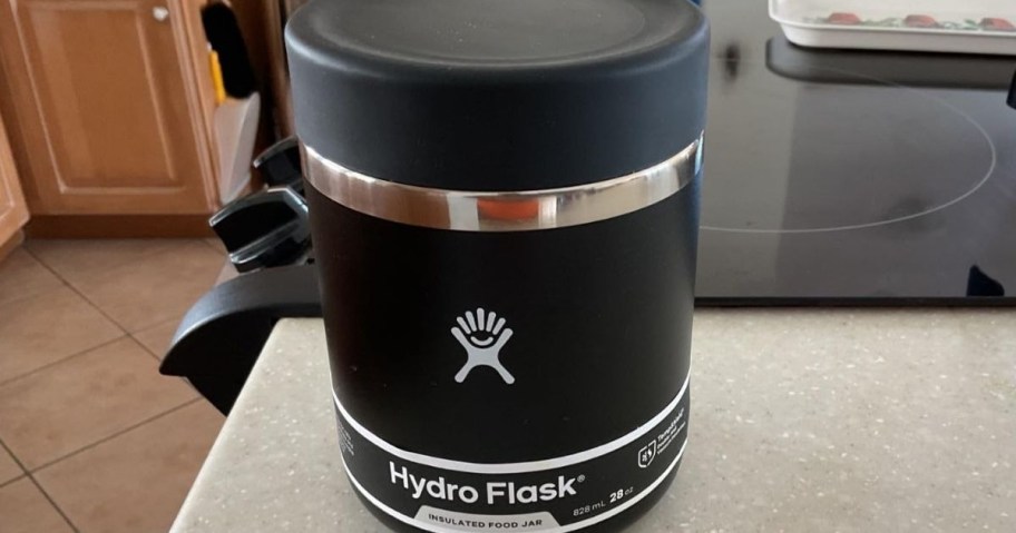 A black Hydro Flask insulated food jar sits on a kitchen countertop near a stove. The jar has a silver accent and a logo.