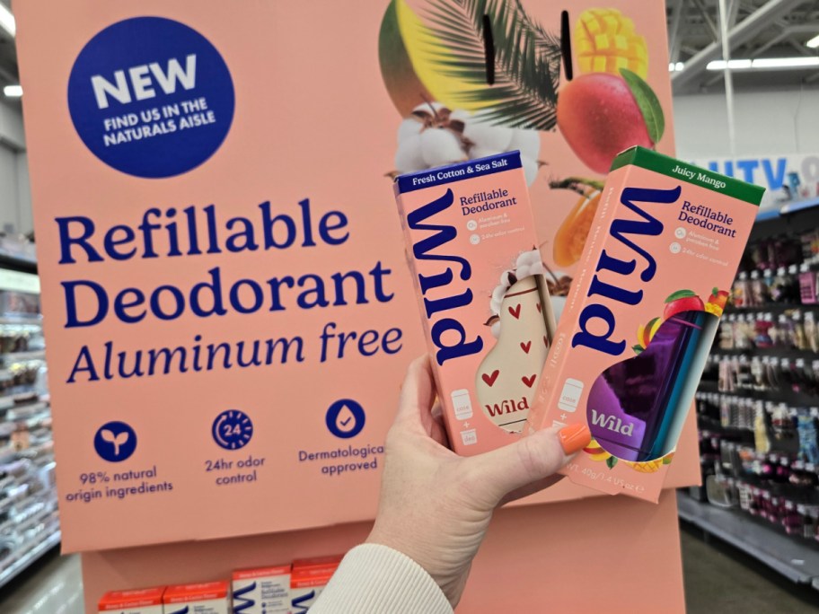 Hand holding two Wild refillable deodorant boxes, Fresh Cotton & Sea Salt, and Juicy Mango. Background displays a sign promoting aluminum-free deodorant.