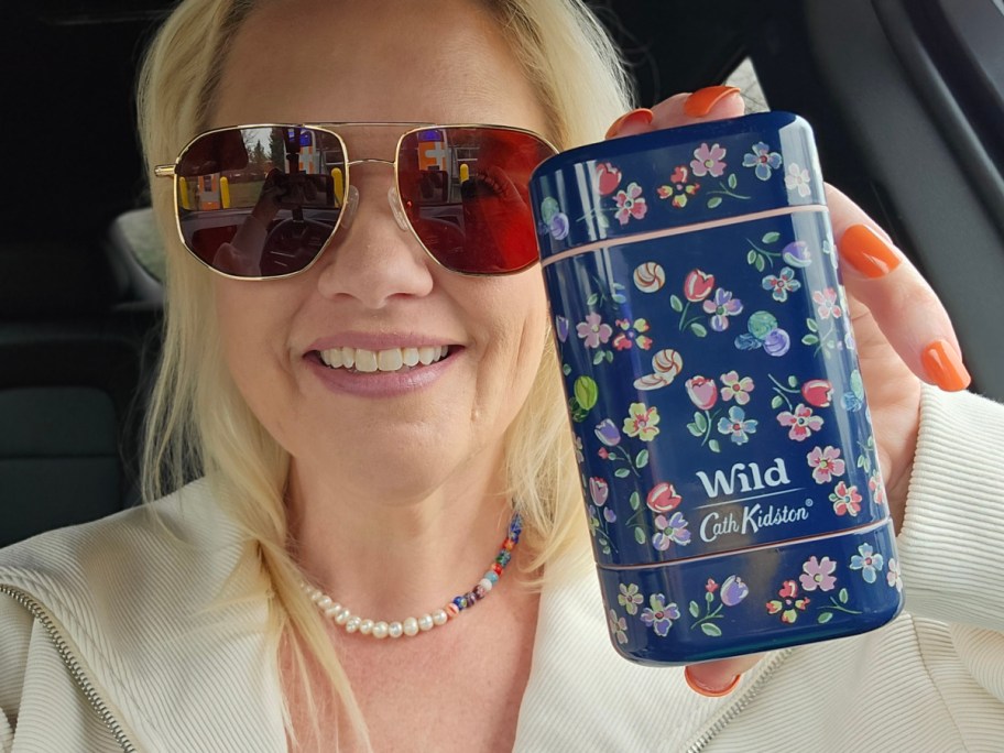 Woman with blonde hair and sunglasses smiles in a car, holding a blue floral-patterned Cath Kidston tin labeled "Wild."