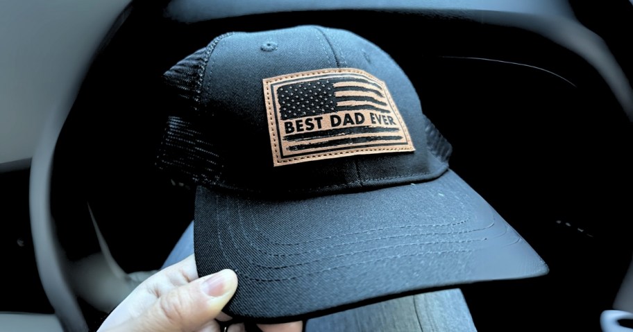 hand holding a black trucker style cap that has a tan badge on it that says "best dad ever"