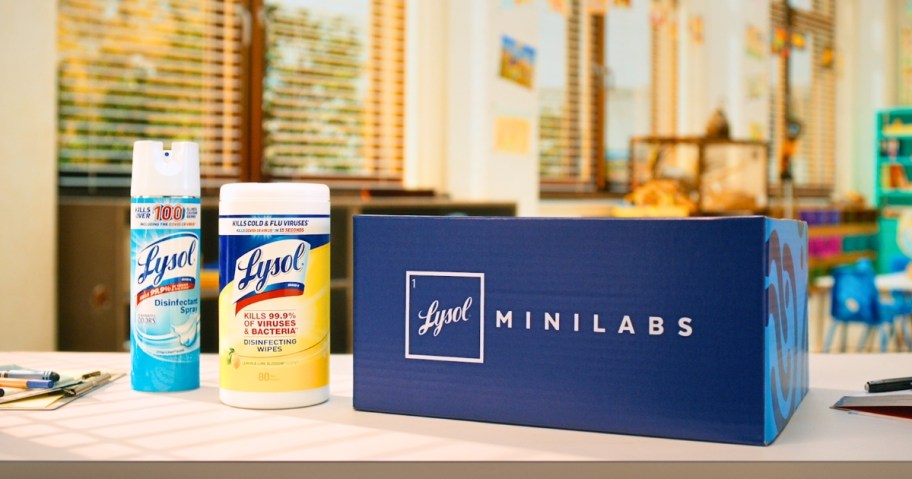 a spray bottle of Lysol and a pack of Lysol wipes next to a Lysol Minilabs Science Kit box on a desk in a classroom