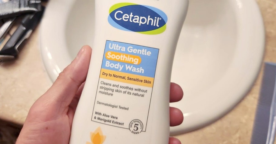 Hand holding a bottle of Cetaphil Ultra Gentle Soothing Body Wash for dry to normal, sensitive skin. It's placed above a white sink.