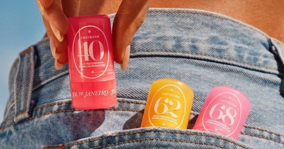 Close-up of three brightly colored perfume bottles, numbered 40, 62, and 68, tucked into the back pocket of faded blue jeans. A hand holds one bottle.
