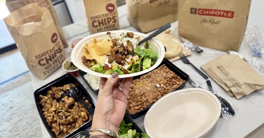 hand holding up a chipotle bowl with trays of ingredients on counter in background