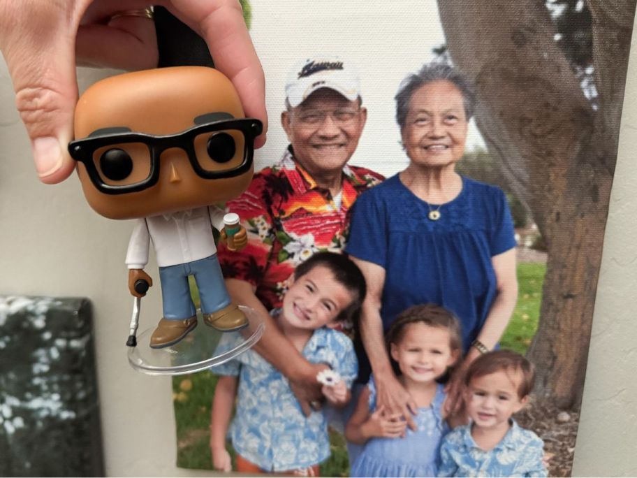 Hand holding a Funko Pop! Me figure next to a family photo