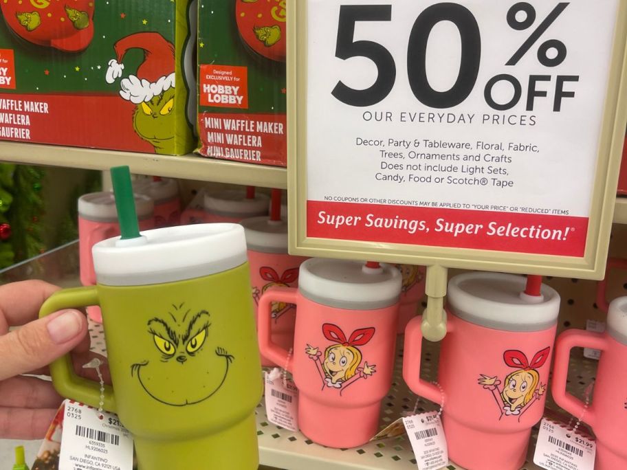 A person holding a green drink tumbler with the Grinch's face on it with pink tumblers in the background