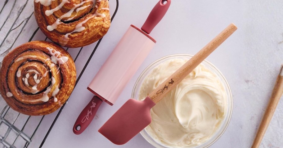 a pink mini rolling pin and spatula by some cinnamon rolls and a bowl of frosting