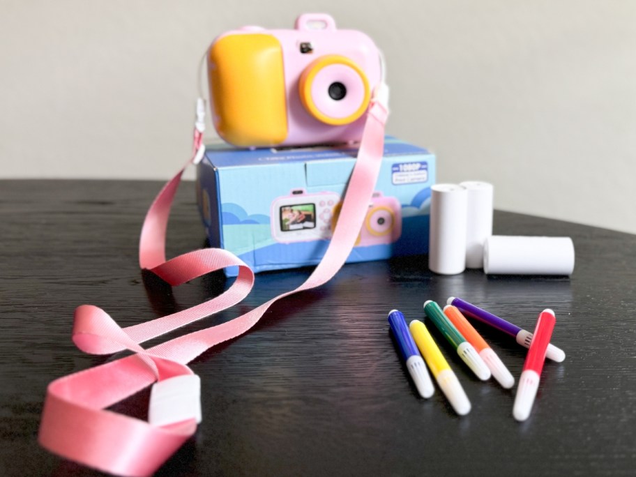 a kids digital camera sitting on top of the box it comes in showing the carry strap, rolls of photo paper and markers