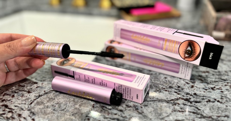 hand holding a mascara wand and 3 boxes with tubes of tarte Tartelette Lash Extension Tubing Mascara on a counter