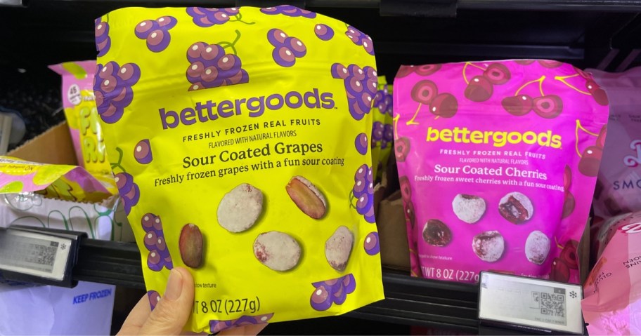 hand holding a bag of bettergoods sour-coated grapes candy next to a bag of sour coated cherries candy