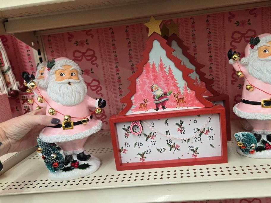 Pink Santa w/ Bells Tabletop D&eacute;cor and Red & White Christmas Tree w/ Santa Countdown Tabletop D&eacute;cor on shelf in store
