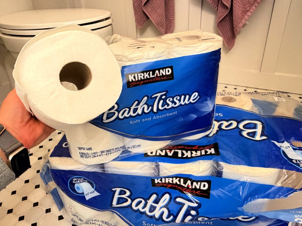 Woman holding up roll of Kirkland bath tissue while standing in bathroom by toilet