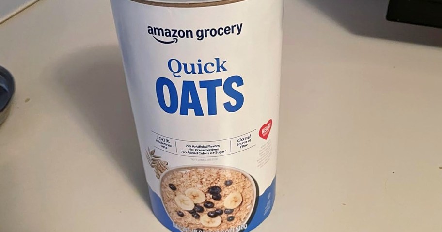 A cylindrical container of Amazon Grocery Quick Oats on a countertop. The label features oats with banana slices and blueberries.