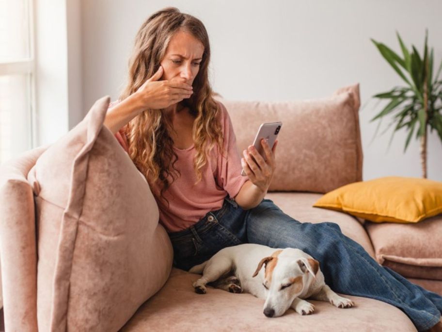 woman with hand over mouth holding cell phone while sitting on couch