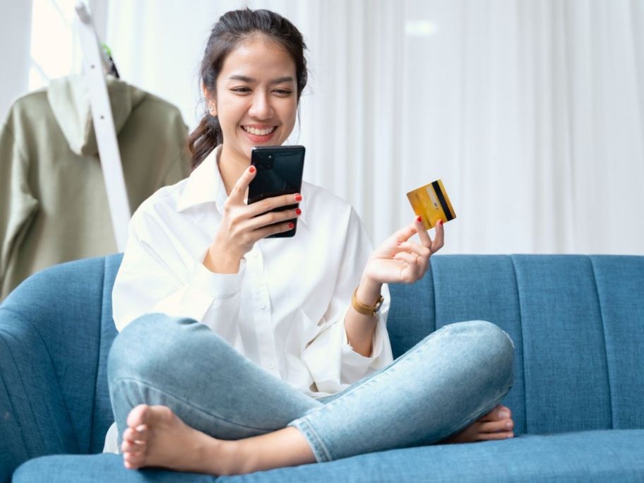 woman sitting on couch and smiling and looking at her cell phone while holding credit card