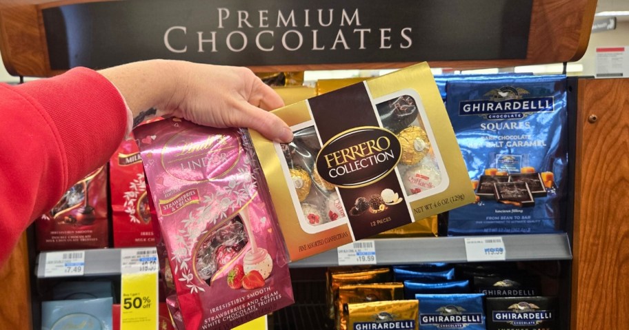 hand holding packs of Lindor Truffles and Ferrero Rocher, bags of Ghirardelli candy on a shelf behind it