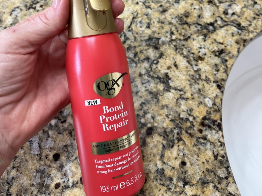 A hand holding a bottle of OGX hair spray.