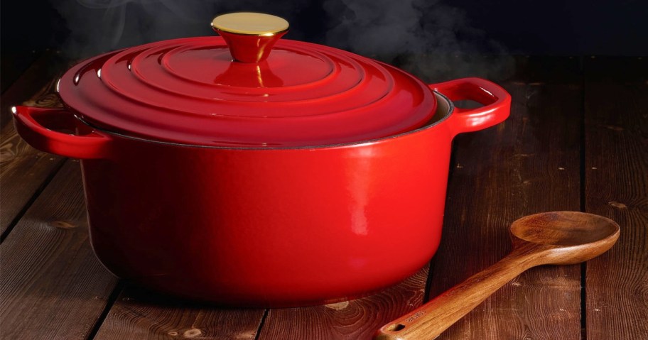 A vibrant red Dutch oven with a golden knob sits on a wooden table, emitting steam. A wooden spoon lies beside it.