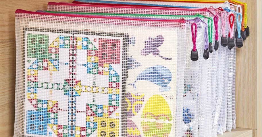 Transparent mesh zipper bags with colorful borders are neatly organized on a shelf. Inside, board games and craft papers are visible.