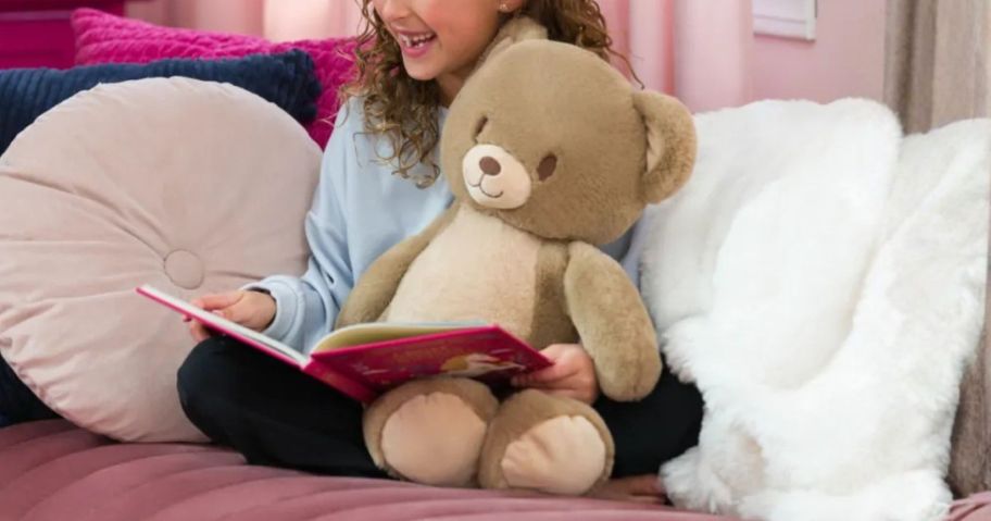 kid holding Heartwarming Hugs brown teddy bear Weighted Stuffed Animal on couch with book