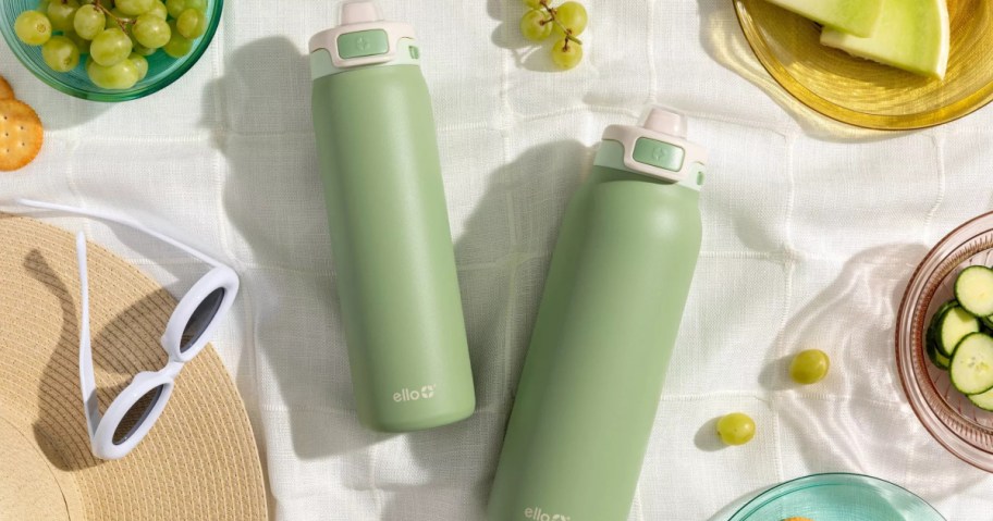 Two green water bottles on a picnic blanket surrounded by grapes, sliced cucumbers, melon, crackers, sunglasses, and a straw hat.